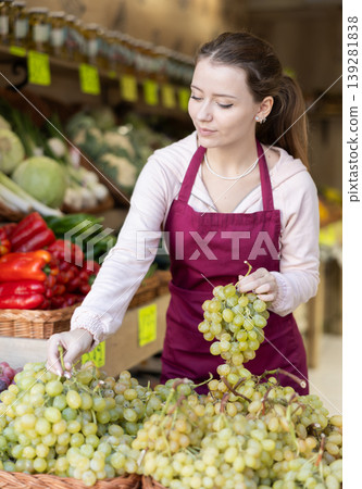 Young woman seller puts grapes in vegetable shop Young woman seller puts grapes in vegetable shop 139281838
