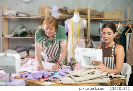Elderly dressmaker making marks on fabric next to young seamstress Elderly dressmaker making marks on fabric next to young seamstress 139281852