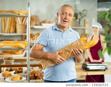 European aged male buyer stands proudly in front of fresh bread holding freshly baked baguettes in bakery store European aged male buyer stands proudly in front of fresh bread holding freshly baked baguettes in bakery store 139282531