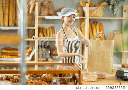 Pretty european young girl seller in apron holding paper bag of baked goods in bakery, ready to meet buyer Pretty european young girl seller in apron holding paper bag of baked goods in bakery, ready to meet buyer 139282535