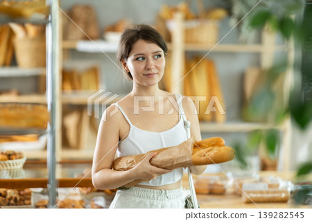 Young female customer stands with baguettes in hands near window of bakery. Young female customer stands with baguettes in hands near window of bakery. 139282545