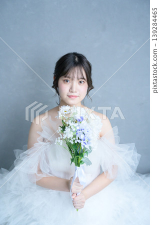 A Japanese woman in a wedding dress gazes out, holding a bouquet against a gray background. 139284465