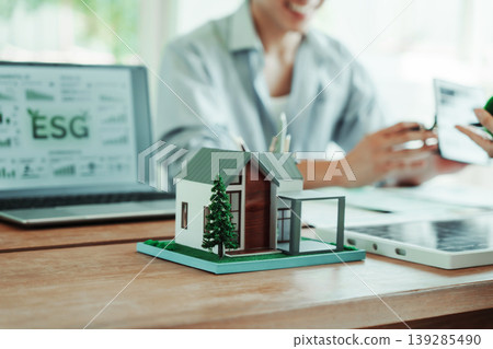 Model house on office desk with laptop showing esg dashboard; colleagues discuss sustainability and green finance in bright modern workspace. 139285490