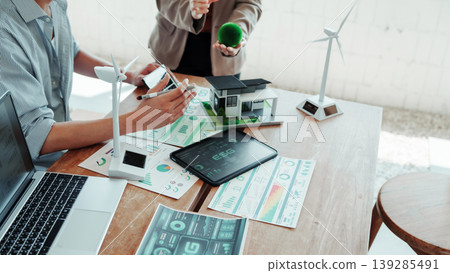 Business team discussing renewable energy project with wind turbine models, solar panels, tablet dashboard, and miniature house on desk with charts and laptop. Business team discussing renewable energy project with wind turbine models, solar panels, tablet dashboard, and miniature house on desk with charts and laptop. 139285491