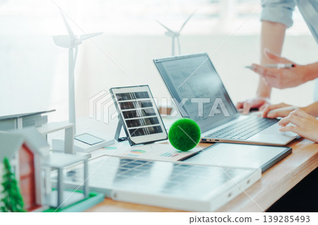 Renewable energy workspace with laptop, solar panel model, wind turbines and eco house mockup on desk, people analyzing data and planning sustainable power project in bright office. Renewable energy workspace with laptop, solar panel model, wind turbines and eco house mockup on desk, people analyzing data and planning sustainable power project in bright office. 139285493