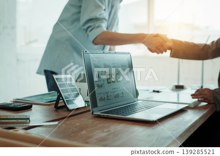Office scene with laptop displaying 'ESG' charts on desk, two people shaking hands in background, tablet and documents on table Office scene with laptop displaying 'ESG' charts on desk, two people shaking hands in background, tablet and documents on table 139285521
