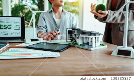Business meeting on sustainable real estate: ESG dashboard on laptop, model eco house on desk, wind turbines and green concept props in modern office. Business meeting on sustainable real estate: ESG dashboard on laptop, model eco house on desk, wind turbines and green concept props in modern office. 139285527