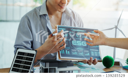 Business team reviews esg metrics on tablet in modern office with renewable energy models, solar panel and wind turbine, highlighting sustainable finance and green strategy. Business team reviews esg metrics on tablet in modern office with renewable energy models, solar panel and wind turbine, highlighting sustainable finance and green strategy. 139285529
