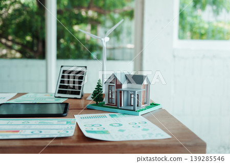 Model house with solar panel and wind turbine on desk with energy charts, sustainable home concept, bright modern office background. Model house with solar panel and wind turbine on desk with energy charts, sustainable home concept, bright modern office background. 139285546