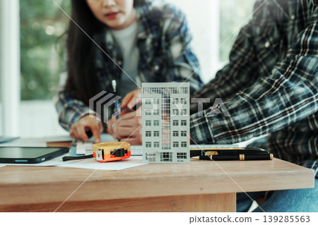 Two people planning building project at table with model apartment, tape measure, and documents, indoors with natural light; home renovation, real estate, architecture teamwork concept. Two people planning building project at table with model apartment, tape measure, and documents, indoors with natural light; home renovation, real estate, architecture teamwork concept. 139285563