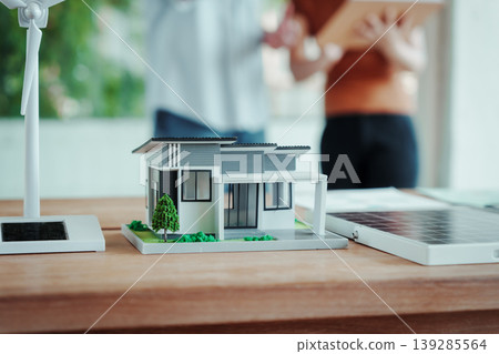 Model house on desk beside solar panel and small wind turbine, with couple discussing in background. Concept of sustainable energy, eco home planning, renewable power. Model house on desk beside solar panel and small wind turbine, with couple discussing in background. Concept of sustainable energy, eco home planning, renewable power. 139285564