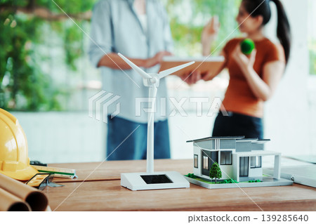 Renewable energy concept: wind turbine model, modern house miniature, and hard hat on desk with architects discussing sustainable design in blurred background. Renewable energy concept: wind turbine model, modern house miniature, and hard hat on desk with architects discussing sustainable design in blurred background. 139285640