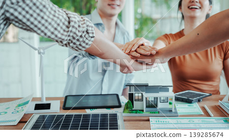 Team stacking hands over desk with solar panel, wind turbine model and eco house mockup in modern office, symbolizing collaboration on renewable energy and sustainable construction. Team stacking hands over desk with solar panel, wind turbine model and eco house mockup in modern office, symbolizing collaboration on renewable energy and sustainable construction. 139285674