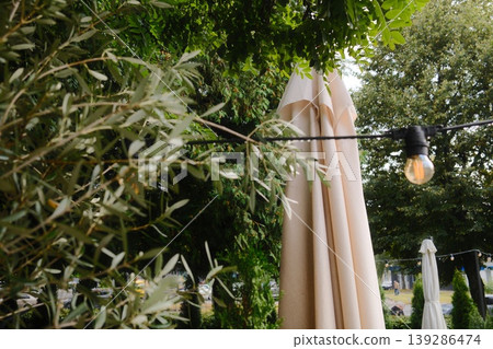 A closed beige umbrella stands next to hanging lights and green trees on a sunny day A closed beige umbrella stands next to hanging lights and green trees on a sunny day 139286474