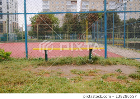 A yellow bench sits in front of a fenced court with buildings in the background A yellow bench sits in front of a fenced court with buildings in the background 139286557