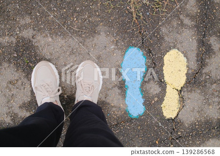 Person wearing shoes stands near blue and yellow painted footprints on asphalt Person wearing shoes stands near blue and yellow painted footprints on asphalt 139286568