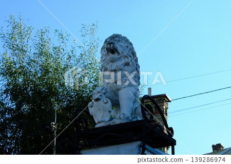 Stone lion statue is sitting on an iron gate, standing guard on a sunny day in a park Stone lion statue is sitting on an iron gate, standing guard on a sunny day in a park 139286974