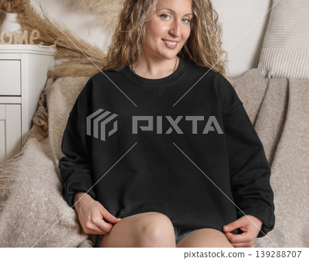 Blank black oversized sweatshirt mockup on fair-haired young woman sitting on sofa, front view 139288707