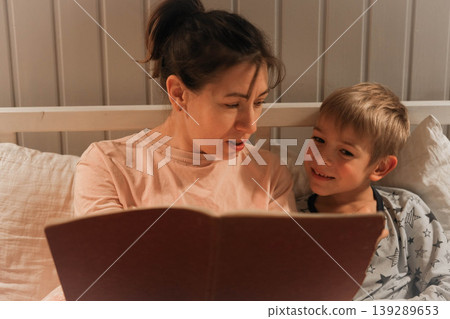 Family bedtime stories. A mother reads a story to her children, wrapped in a blanket. Children listen to soothing stories. A cozy family scene before bed in a softly lit bedroom. Copy space. Banner 139289653