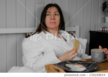 A calm woman enjoys a quiet breakfast. A serene lady sits in bed in a robe, holding a mug and a cracker, in the cozy atmosphere of her home. Morning rituals alone. A cozy interior. Copy space. Banner A calm woman enjoys a quiet breakfast. A serene lady sits in bed in a robe, holding a mug and a cracker, in the cozy atmosphere of her home. Morning rituals alone. A cozy interior. Copy space. Banner 139289654