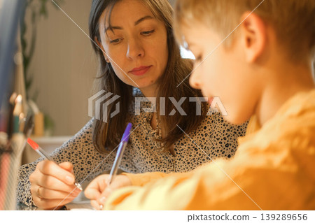 A mother helping her son with his studies. Homework assistance. Warm lighting. A cozy atmosphere for family learning. A focused student's pose. A scene of studying before bed. Space for text. Banner. 139289656