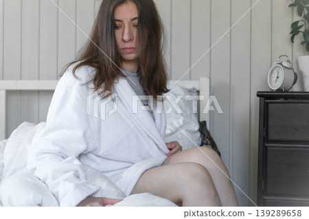 Woman in white robe on bed, disheveled hair, soft sunlight through window, clock on nightstand showing morning time, pensive freelancer getting ready for a new day, neutral tones in bedroom. Banner 139289658
