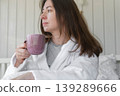 A woman lies on a pastel-colored bed, holding a coffee mug. A calm woman in a robe enjoys the serene morning atmosphere at home. Morning routine. Self-care. Starting a new day. Space for text. Banner 139289666