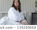 Woman in white robe on bed, disheveled hair, soft sunlight through window, clock on nightstand showing morning time, pensive freelancer getting ready for a new day, neutral tones in bedroom. Banner 139289668