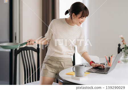 Happy young asian woman holding frying pan while working on laptop for business checking smartphone app with cheerful smile standing in bright home kitchen preparing food Happy young asian woman holding frying pan while working on laptop for business checking smartphone app with cheerful smile standing in bright home kitchen preparing food 139290029