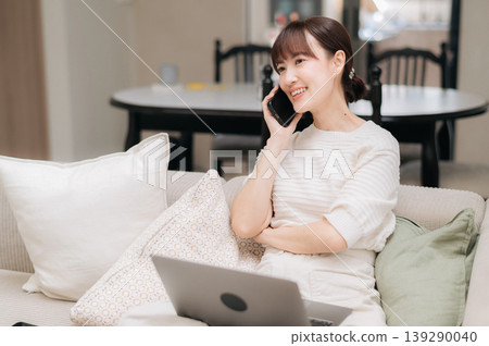 Happy young asian woman enjoying phone call about business app on smartphone while using laptop computer relaxing on comfortable sofa inside bright modern home living room Happy young asian woman enjoying phone call about business app on smartphone while using laptop computer relaxing on comfortable sofa inside bright modern home living room 139290040