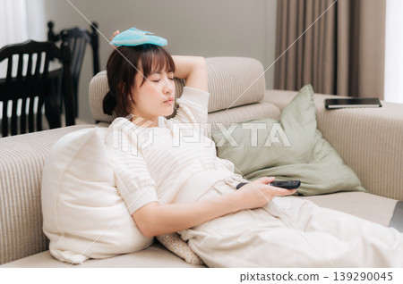Sick young asian woman with cooling pad on head resting on comfortable sofa feeling tired and unwell holding remote control away from business app on smartphone and closed laptop Sick young asian woman with cooling pad on head resting on comfortable sofa feeling tired and unwell holding remote control away from business app on smartphone and closed laptop 139290045