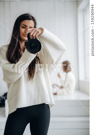 Professional female photographer is standing indoors with camera, kid sitting at background, behind 139290944