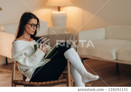 Smart person, in glasses. Relaxed woman is sitting in the chair and reading the book at home Smart person, in glasses. Relaxed woman is sitting in the chair and reading the book at home 139291131