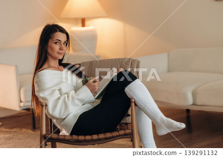 Cozy room, feeling good. Relaxed woman is sitting in the chair and reading the book at home Cozy room, feeling good. Relaxed woman is sitting in the chair and reading the book at home 139291132