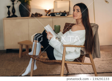 Cozy room, feeling good. Relaxed woman is sitting in the chair and reading the book at home Cozy room, feeling good. Relaxed woman is sitting in the chair and reading the book at home 139291133