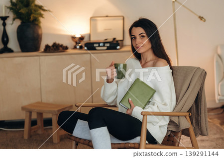 Beautiful brunette is with cup of tea. Relaxed woman is sitting in the chair and reading the book at home 139291144