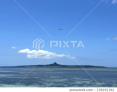Ie Island as seen over the rocky reefs of Bise Coast, with an aircraft flying overhead. Ie Island as seen over the rocky reefs of Bise Coast, with an aircraft flying overhead. 139291162