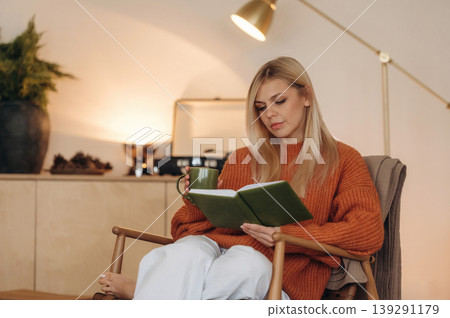 With cup of tea. A woman is sitting on the chair in the room and reading the book 139291179