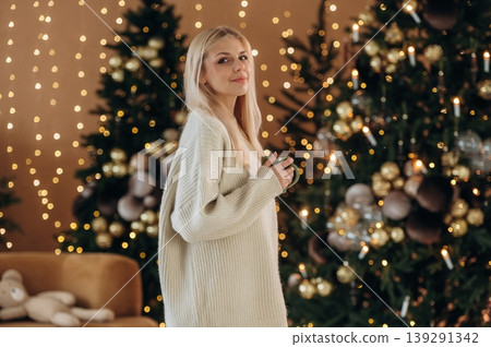 Standing and posing. Beautiful woman is standing in the room with New Year garlands and fir Standing and posing. Beautiful woman is standing in the room with New Year garlands and fir 139291342