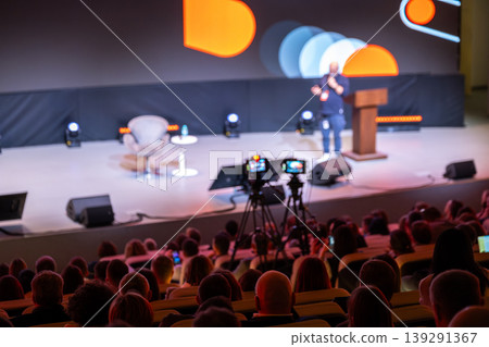 Conference speaker presenting on stage to large audience with cameras capturing the event Conference speaker presenting on stage to large audience with cameras capturing the event 139291367