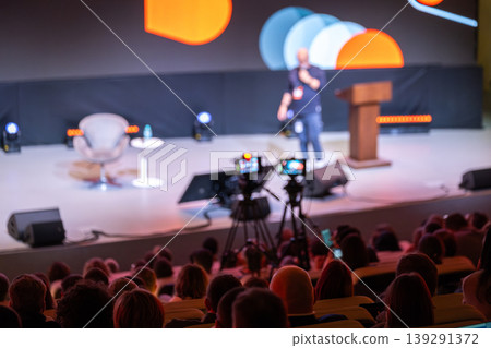 Conference speaker on stage with audience and cameras filming live presentation at event Conference speaker on stage with audience and cameras filming live presentation at event 139291372