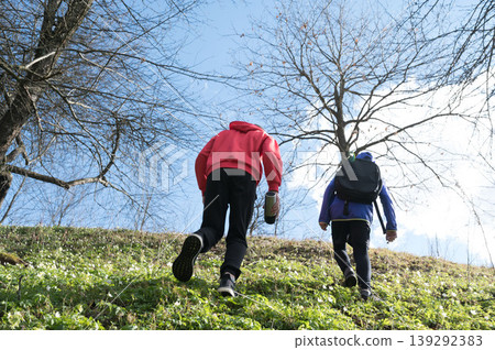 Two hikers ascend a grassy hill under a bright sky, surrounded by leafless trees. Two hikers ascend a grassy hill under a bright sky, surrounded by leafless trees. 139292383