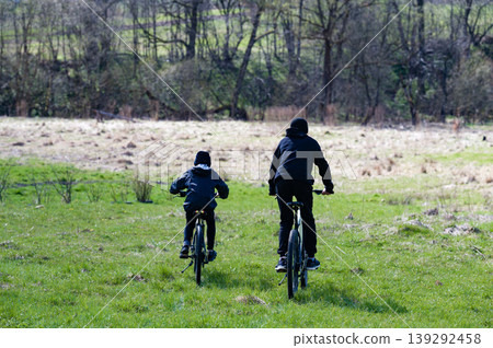 Two cyclists ride through a verdant field, trees forming a backdrop. A peaceful outdoor scene. 139292458