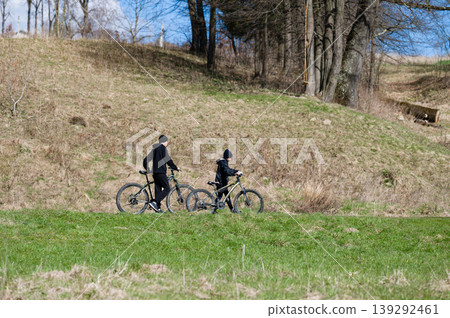 Two cyclists enjoying a sunny day, biking along a grassy path. A scenic woodland backdrop adds to the peaceful atmosphere. 139292461