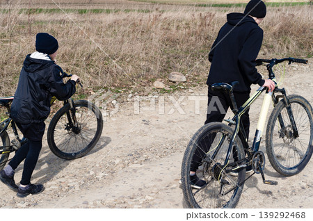 Two kids walking their bikes down a dirt road. A sunny day outdoors. 139292468