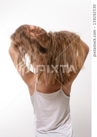 Girl with long brown hair, viewed from behind, holding her hair. She's wearing a white tank top against a white background. Girl with long brown hair, viewed from behind, holding her hair. She's wearing a white tank top against a white background. 139292530