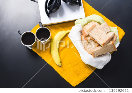 Simple breakfast with coffee, bread, banana, book and headphones on a black surface. Simple breakfast with coffee, bread, banana, book and headphones on a black surface. 139292651