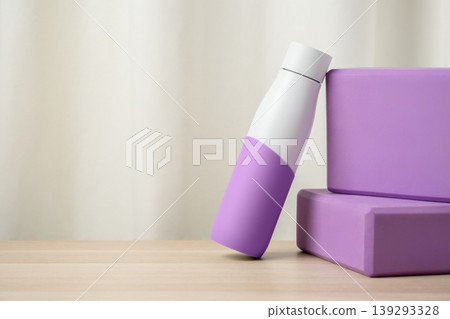 Purple insulated water bottle leaning against two matching yoga blocks on a wooden surface, evoking fitness, hydration, yoga practice and a calming, minimalist wellness studio atmosphere. Purple insulated water bottle leaning against two matching yoga blocks on a wooden surface, evoking fitness, hydration, yoga practice and a calming, minimalist wellness studio atmosphere. 139293328