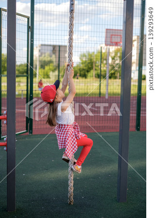 A little preschool-age girl climbs up a rope on a playground in the yard. 139293649
