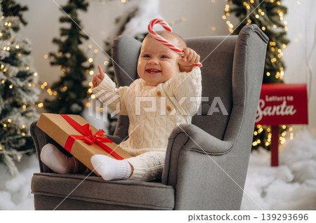 Sweet candy in hand. Little baby is sitting indoors in the Christmas decorated room Sweet candy in hand. Little baby is sitting indoors in the Christmas decorated room 139293696